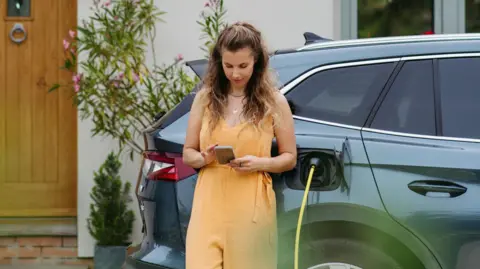Getty Images Woman wearing a peach-coloured sleeveless jumpsuit looking down at her smartphone while leaning backwards against an electric SUV car, which has a yellow charging leading plugged into it. Behind is the front of a cottage-style house.