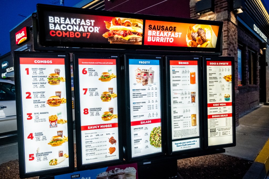 Wendy's drive-thru menu with Breakfast Baconator Combo, Sausage Breakfast Burrito, various combos, Frosty desserts, drinks, sides, and sweets displayed on illuminated screens at night.