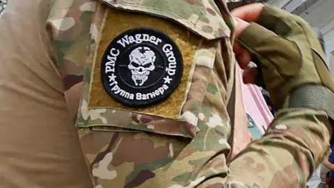 Reuters A close-up of a gloved hand and arm of a Wagner soldier, showing his camouflaged fatigues and the black and white Wagner logo featuring a white skull