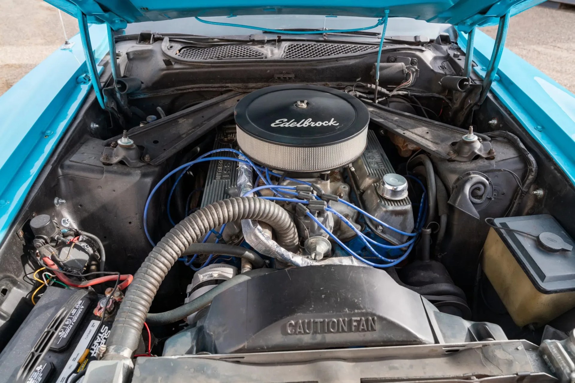Car engine bay with an Edelbrock air filter and blue ignition wires inside a turquoise vehicle.
