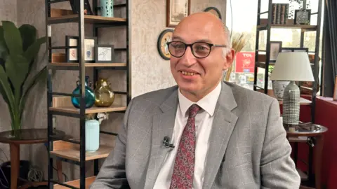 Dr Tarek Tawfik wears a grey suit, a red tie and glasses. He smiles at a slight angle to the camera. Behind him, his office is filled with stand-alone shelves holding vases, books and picture frames. 