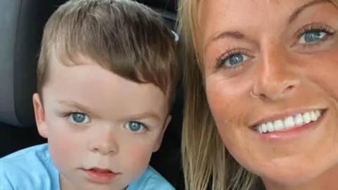 Tammy McDaid A little boy with short light brown hair and blue eyes is looking at the camera while sitting in a car with his mum. His mum has blue eyes and long blonde hair and is wearing a nose ring. She is smiling