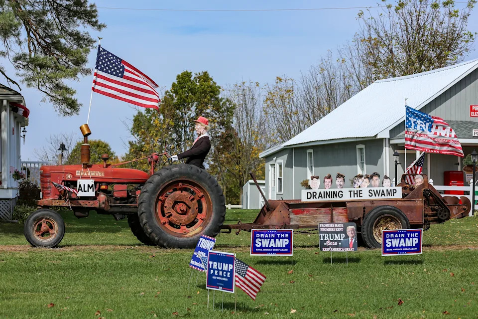 Man on tractor with U.S. flag, pulls a cart with "Draining the Swamp" display and political signs on a grassy lawn next to a house