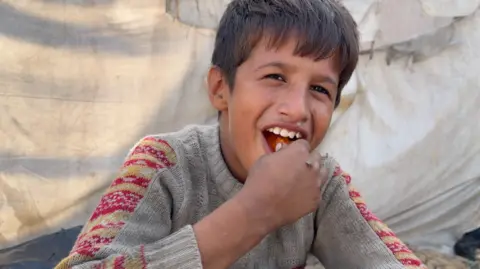 A boy, with medium-length dark hair, bites into some food in his hand. He looks happy with his mouth open wide. Behind him is what looks like a white canvas tent and he is wearing a grey knitted jumper with yellow and red decoration on the tops of the sleeves.