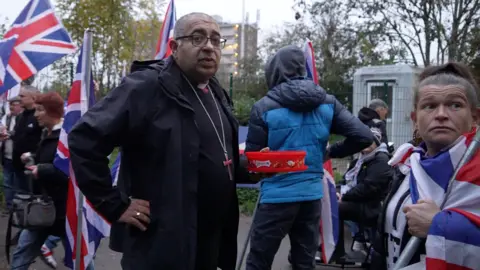 BBC A bishop in black religious clothing stands with his hand on his hip looking at the camera holding an open red tub of Celebrations chocolates, surrounded by protesters draped in Union Jacks.
