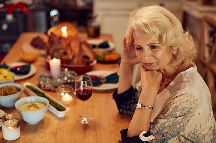 Older woman sitting alone at Thanksgiving dinner table looking sad while in-laws make fun of her holiday meal.