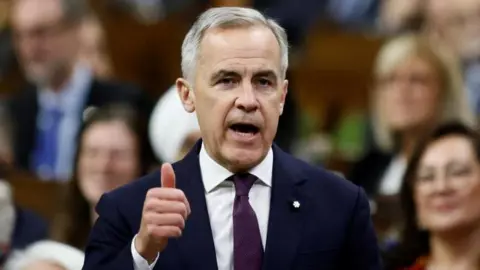 Reuters Canada's Prime Minister Mark Carney speaks during Question Period in the House of Commons on Parliament Hill in Ottawa, Ontario, Canada. He is wearing a navy blue suit and a dark purple tie, and is gesturing with a thumbs-up mid-speech as he addresses the House of Commons. 