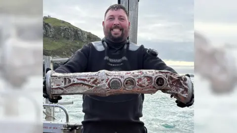 David Kennard/Neptune's Army of Rubbish Cleaners A man in a black wetsuit smiles as he holds a cylindrical metal object believed to be a sonobuoy. He is stood on a boat with, the sea and a portion of land can be seen behind him. He has short brown hair and a brown goatee style beard. 