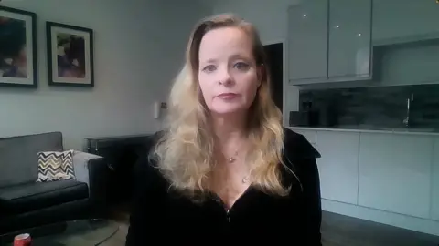 BBC Dayna Arnold she has shoulder length blond hair and is wearing a black top and is sat in front of units in her kitchen