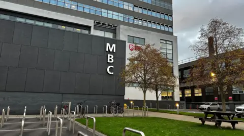 The front of a large grey building with lots of windows. MBC is written on the nearest wall which is a darker shade of grey and does not have windows. The Queen's University Belfast emblem can be seen through the leaves of a tree in the forecourt. There are a few bicycles among the bike stands out front. There are also pathways between grass in the forecourt.