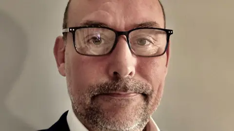 Stephen Walton A closeup of Stephen who has very short cropped brown hair, which is receding from the forehead, and a thin beard. He is wearing square black-framed specs on his face and a dark suit. 