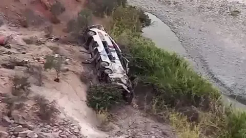 Reuters A frame from a video showing a wrecked passenger bus at the bottom of a deep ravine. The terrain is rocky and sandy with a shallow river snaking across the bottom of the ravine. The bus looks utterly destroyed.