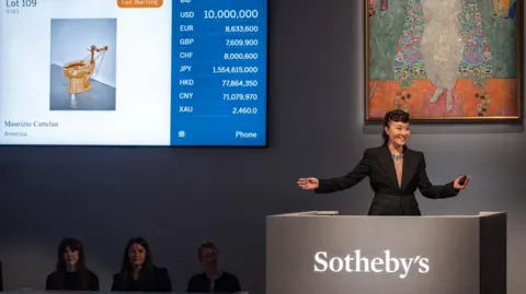 Sotheby's A woman in a smart black suit at a podium in the auction house. A picture of the gold toilet is on a screen. A Gustav Klimt artwork is also on show.