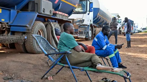 AFP/Getty Drivers of Malian tanker trucks sit on chairs beside their vehicles - one dressed in blue overalls checks his mobile phone - as they wait to cross the border between Ivory Coast and Mali - 31 October 2025.