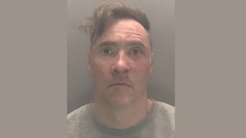 CPS A mugshot of Paul Doyle wearing a grew jumper looking stunned. His hair, a long brown fringe, is dishevelled and to the side. He is wearing a grey t-shirt.