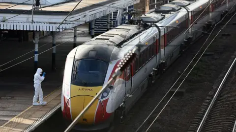 Reuters The red, yellow and grey LNER train standing at a platform in Huntingdon station. A forensic officer with white overalls stands on the platform at the front of the train. 