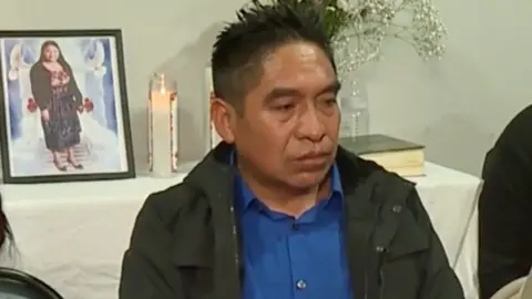 CBS Mauricio Velazquez in a dark coat and blue button-up shirt. Behind him is a photograph of his late wife, Maria Florinda Rios Perez, flowers and a candle. 