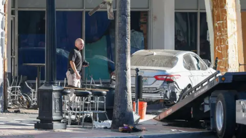 Tampa Bay Times Image of an official standing beside a wrecked car, next to a tow truck, in front of a bar with overturned bar stools