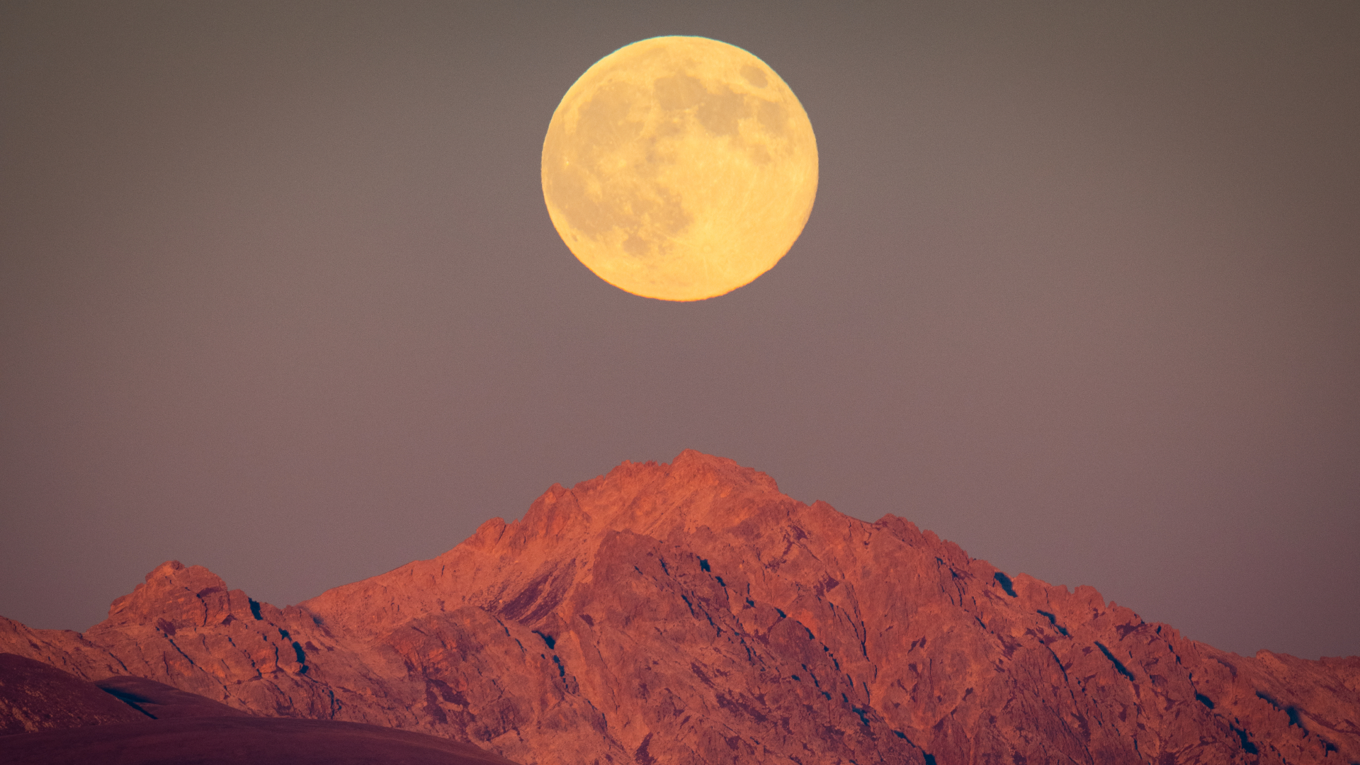 A yellow full moon is pictured rising above a rocky mountain against a pale grey-blue sky.