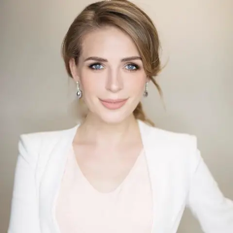 National Crime Agency A woman smiling with her hair tied back and wearing a smart white jacket over a pale pink top. 