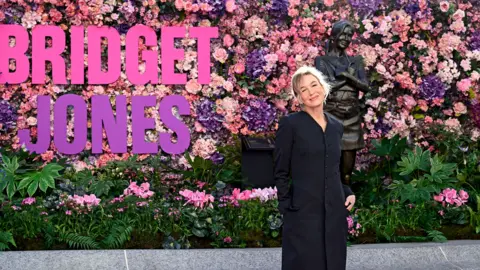 Getty Images Renée Zellweger smiles while wearing a black dress and posing next to a statue of Bridget Jones next to a sign that says 'Bridget Jones' against a backdrop of pink and purple flowers lining the wall, in Leicester Square in London on Monday.