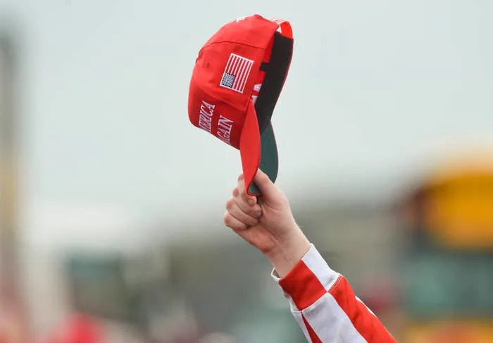 Person holding up a red cap with "Make America Great Again" text and an American flag patch at a public event