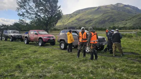 Carabineros de Chile/Reuters Three large vehicles parked on a grassy field. A group of people are standing next to the cars. They are wearing hi-vis safety gear. A rocky hillside is in the distance behind them.