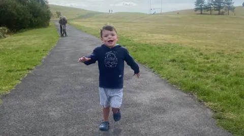 Tammy McDaid A young boy is smiling while running along a path in the countryside. He is wearing a blue hooded top and blue and white striped shorts and blue croc shoes. There are shrubs and trees in the distance. 