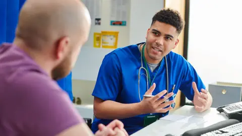 Getty Images A young male doctor is talking to a male patient in a doctors office. We can see the doctors face but only the back of the patients head. The doctor is in blue scrubs and the patient is wearing a purple T -shirt