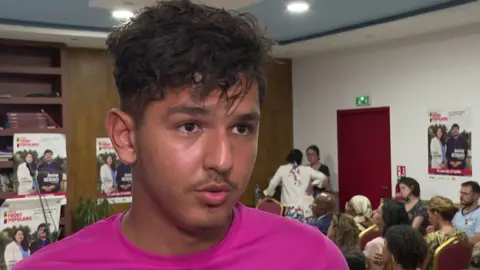 AFP via Getty Images Mehdi Kessaci is speaking to someone off camera in a room where there are many posters of his brother in the background. Mehdi is wearing a bright pink t-shirt and has medium length dark hair and a goatie.