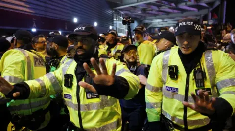 Reuters A group of male officers gather outside the stadium. Two of them, one a black man with a moustache and the other white, both wearing caps, have their hands outstretched pushing people back 