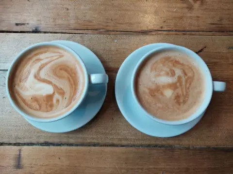 Grace Dean/BBC Two cups of coffee, in white cups on white saucers, pictured on a brown wooden table