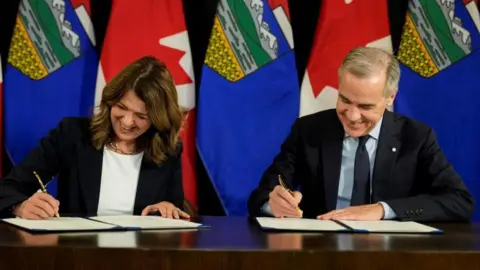 Reuters Canada's Prime Minister Mark Carney and Alberta's Premier Danielle Smith sign a Memorandum of Understanding (MOU), before the Prime Minister's energy-related announcement in Calgary, Alberta, Canada November 27, 2025. They are seated, and pictured each signing a document. Behind them is a row of Canada and Alberta flags. 