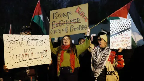 Reuters Protesters hold signs outside the stadium before the match. They have Palestinian flags. Two signs say "boycotts not bullets" and "take the foot off their necks and get off our pitch".