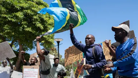 AFP via Getty Images Protesters waving a Tanzanian flag gesture while holding placards 