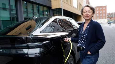Barry Cronin Ford UK's managing director Lisa Brankin wearing blue jeans, a navy blue blouse with white polka dots and a navy blue cardigan. She is standing next to a black Ford Capri connecting it to an electric charger and smiling at the camera