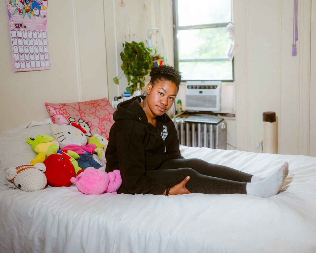 Adriana Del Orden sits on her bed in a bright bedroom decorated with stuffed animals and a pink Hello Kitty pillow. Sunlight streams through a window behind her.