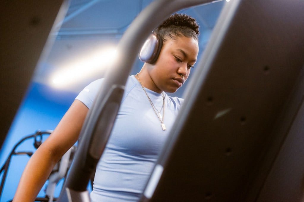 Adriana Del Orden walks on a treadmill at a gym, wearing headphones and a light blue shirt. The shot is framed through exercise equipment.