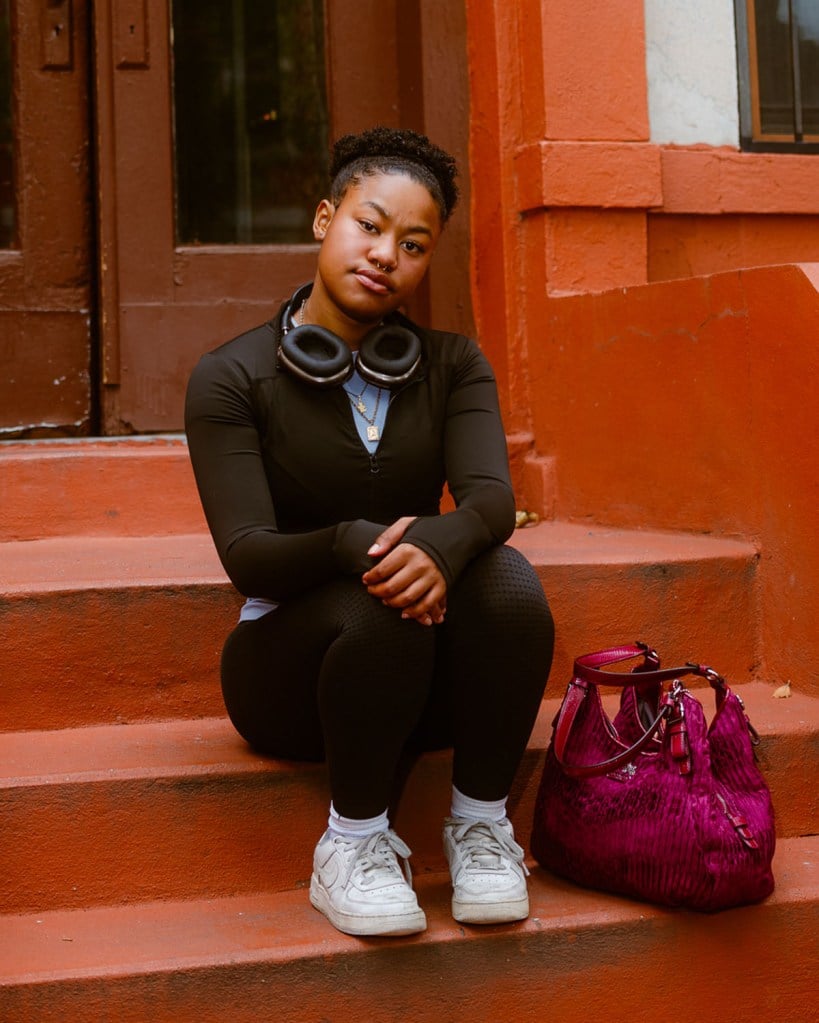 Adriana Del Orden sits on the steps of a brownstone in New York City, wearing black athletic clothes with a pair of headphones around her neck and a bright magenta bag beside her.