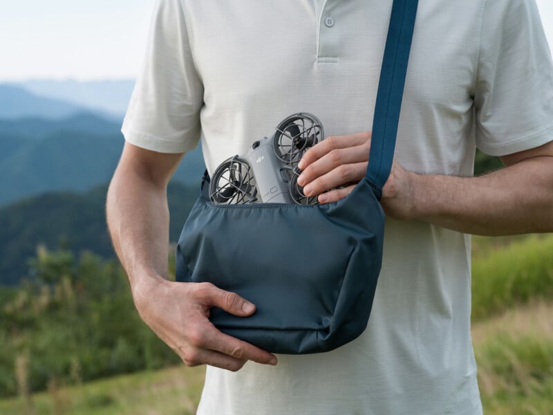 A person in a white shirt holds a blue shoulder bag with a small drone partially visible inside, standing outdoors with green hills and mountains in the background.
