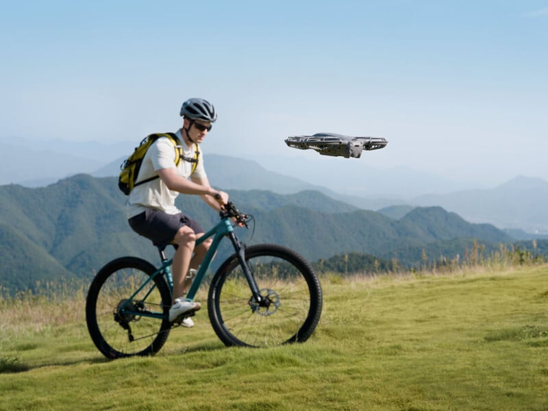 A person wearing a helmet and backpack rides a mountain bike on a grassy hill with mountains in the background, while a drone hovers nearby in the clear sky.