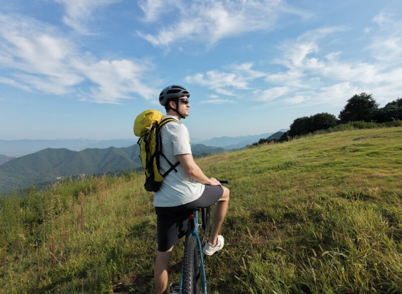 A person wearing a helmet and yellow backpack stands with a bicycle on a grassy hill, gazing at distant mountains under a blue sky with scattered clouds.