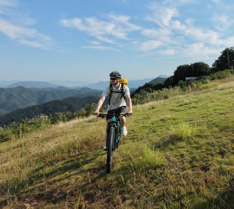 A person wearing a helmet and backpack rides a mountain bike uphill on a grassy slope, with green hills and mountains in the background under a blue sky with wispy clouds.