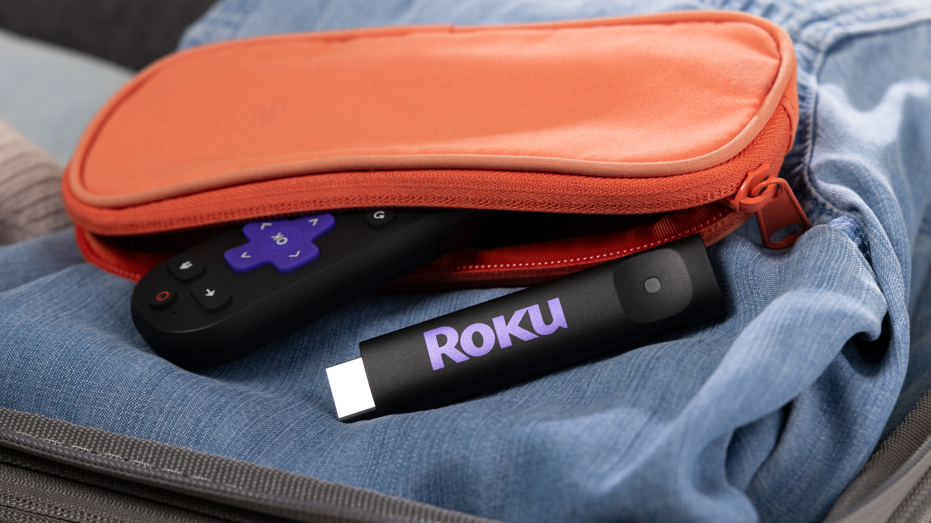The 2025 Roku Streaming Stick and a Roku remote control emerging from an orange-colored pouch, all of which are sitting atop folded denim