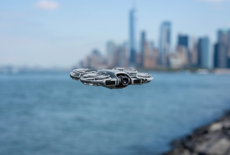 A small drone hovers above the water with a blurred city skyline in the background on a clear day. The drone is in sharp focus, showing its camera and propeller guards.
