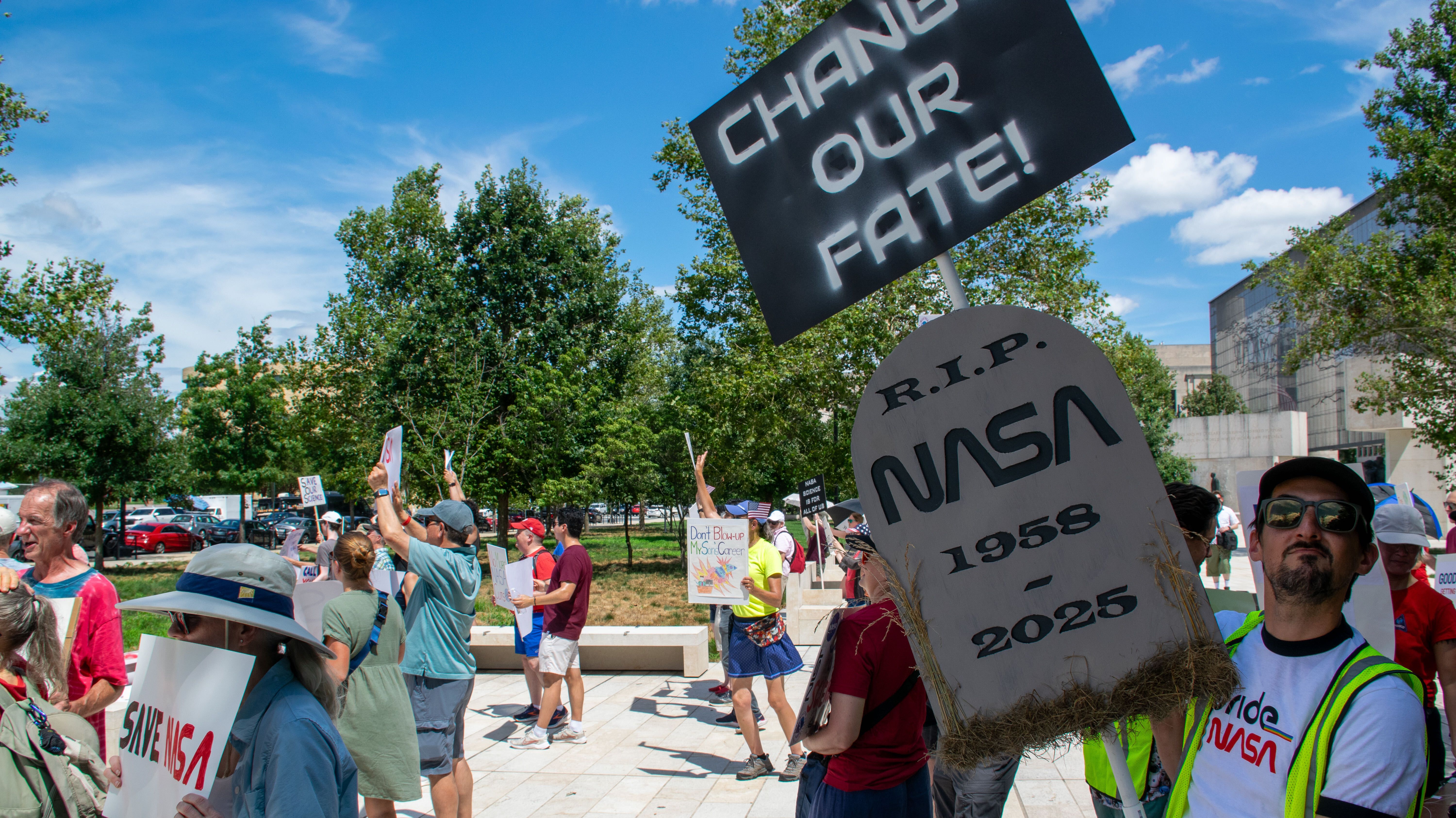 People hold signs outside on a sunny day in protest of NASA budget cuts, July 20, 2025.