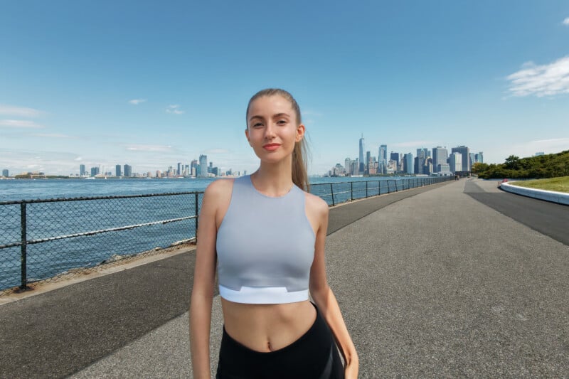 A woman in athletic wear stands on a paved path by the water, with a city skyline in the background under a clear blue sky. She is smiling and appears to be outdoors on a sunny day.