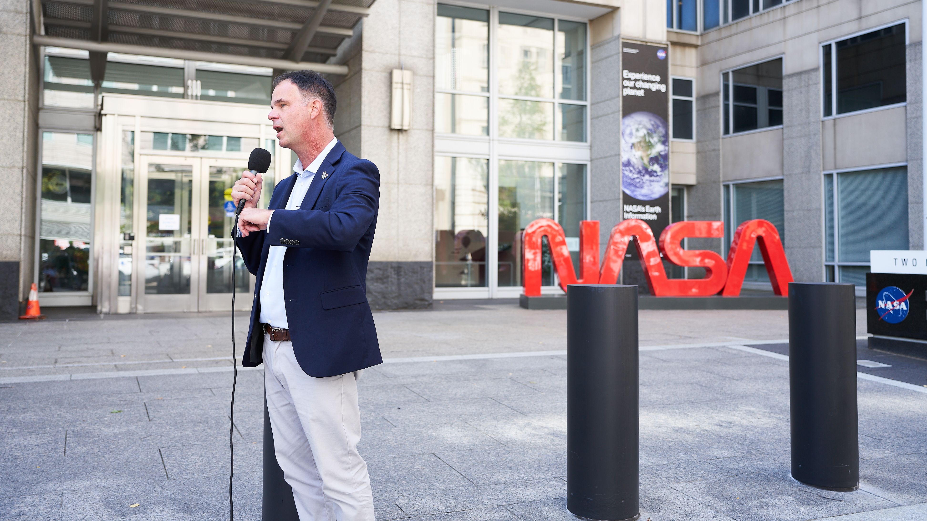 A man stands outside holding a microphone in front of a NASA sign.