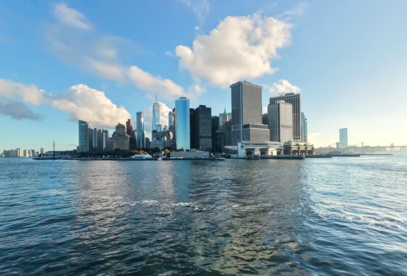 A view of the New York City skyline seen from across the water, with tall skyscrapers, blue sky, scattered clouds, and reflections shimmering on the surface of the river.