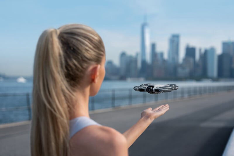 A woman with blonde hair in a ponytail holds out her hand as a small drone hovers above it, with a city skyline and waterfront visible in the background.